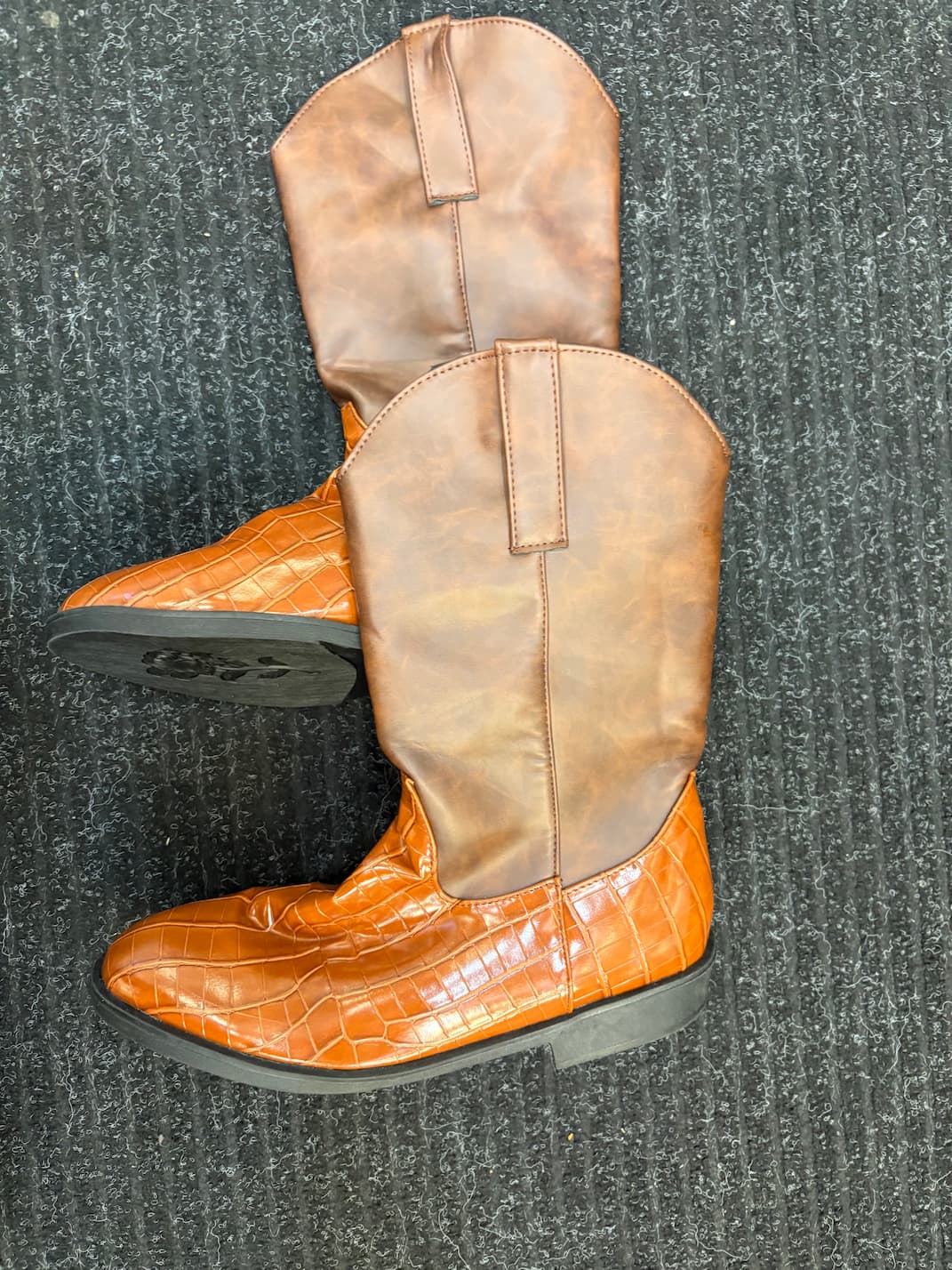 A pair of cowboy boots with tan faux-leather shafts and bright orange crocodile-embossed toe caps, displayed on a dark gray textured carpet.