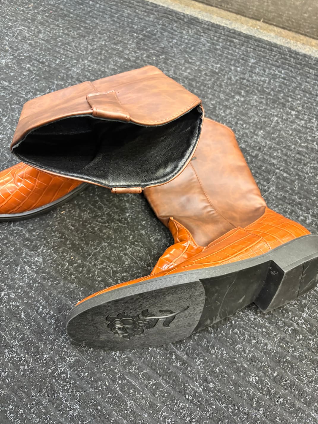 A pair of tall brown leather boots with a glossy crocodile-embossed orange lower section and black soles, shown on a gray textured carpet.