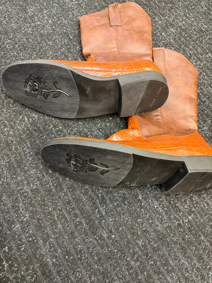 A pair of cowboy boots with bright orange crocodile-embossed leather shafts and tan leather lower sections, shown from the bottom to display the soles with a floral engraving, placed on a gray textured carpet.