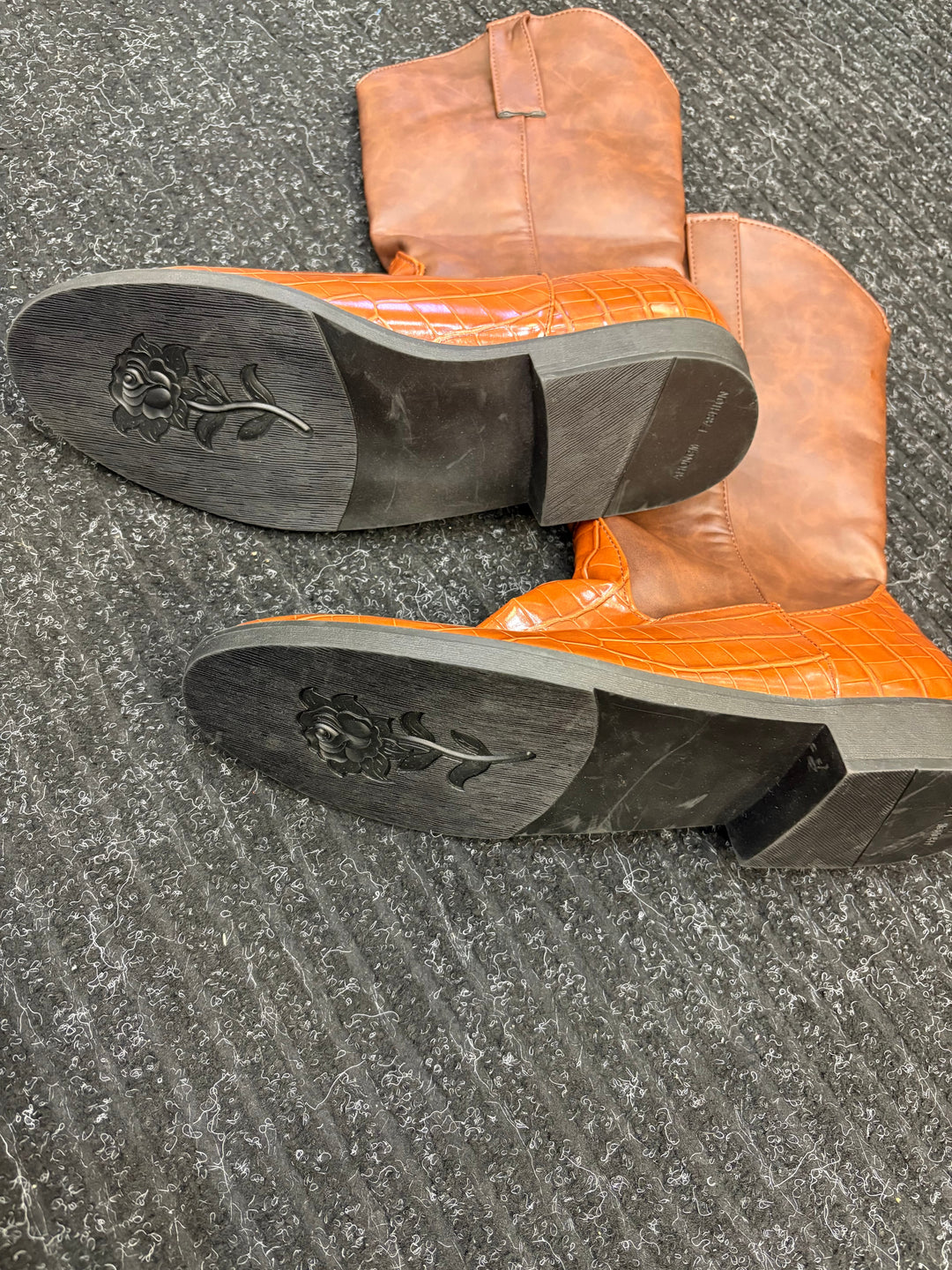 A pair of cowboy boots with bright orange crocodile-embossed leather shafts and tan leather lower sections, shown from the bottom to display the soles with a floral engraving, placed on a gray textured carpet.