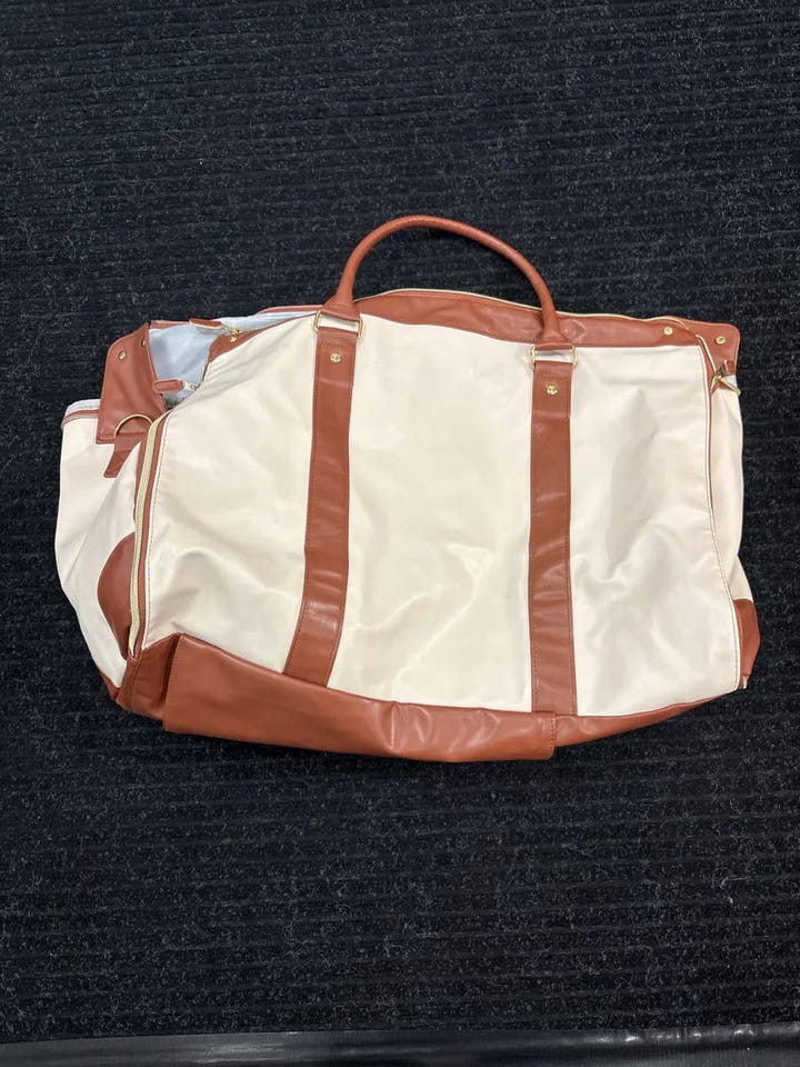 A cream and brown duffel bag with a top handle, laid flat on a dark textured carpet.