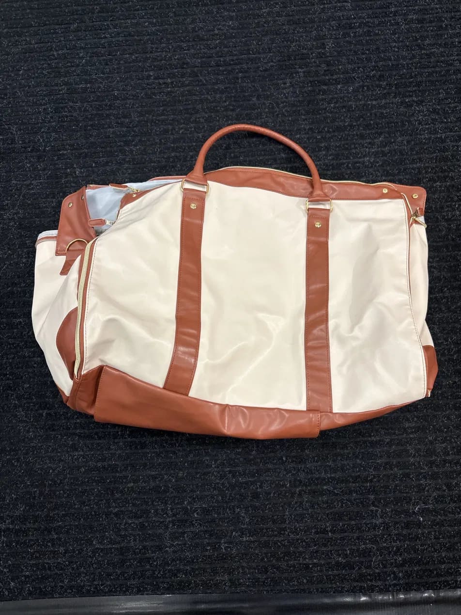 A cream and brown duffel bag with a top handle, laid flat on a dark textured carpet.