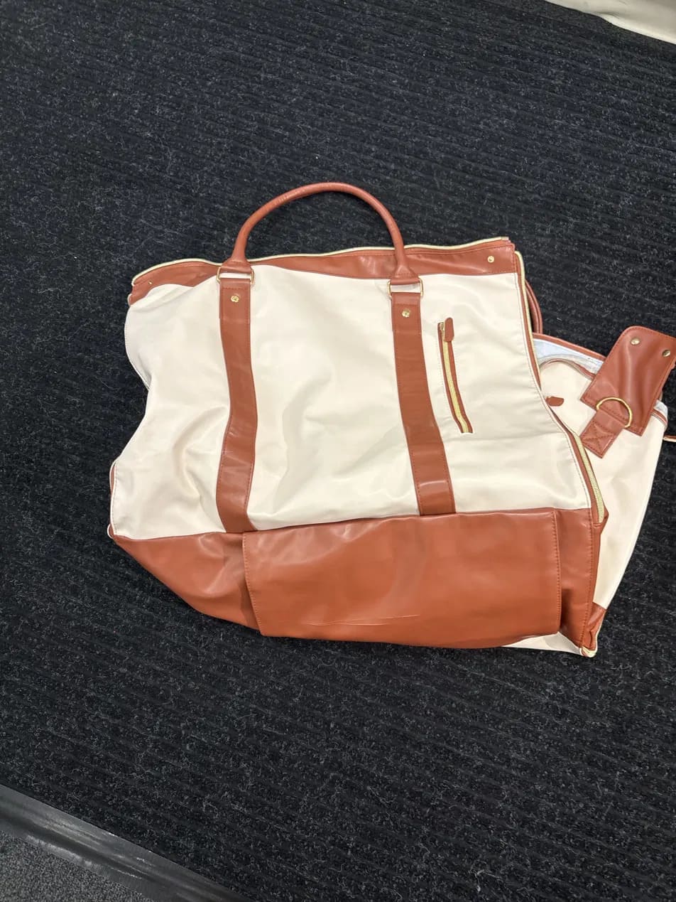 A large tote bag with cream canvas upper panels and smooth tan-brown leather trim, handles, and base, photographed on a dark textured carpet.