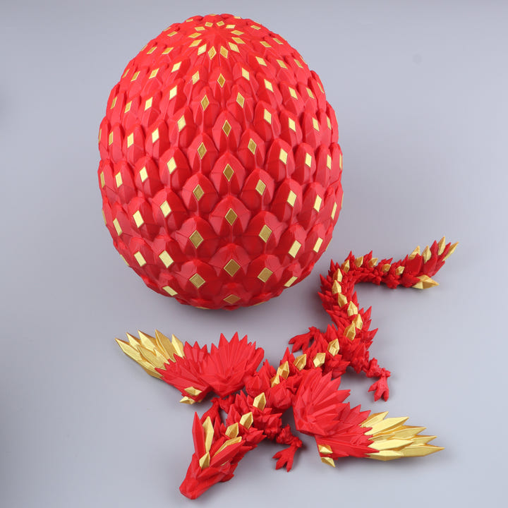 A red dragon figurine with gold accents lies in front of a large red egg-shaped structure covered in repeating gold diamond patterns, photographed on a light gray studio background.