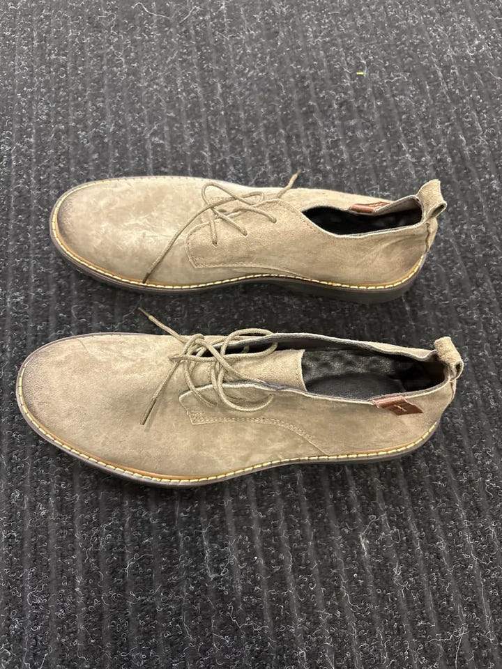 A pair of light beige suede lace-up shoes with a low-cut vamp, displayed on a dark gray ribbed carpet.