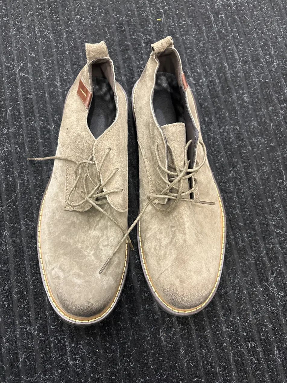 A pair of light beige suede lace-up boots with light tan stitching along the sole, laid flat on a dark gray ribbed textured surface.