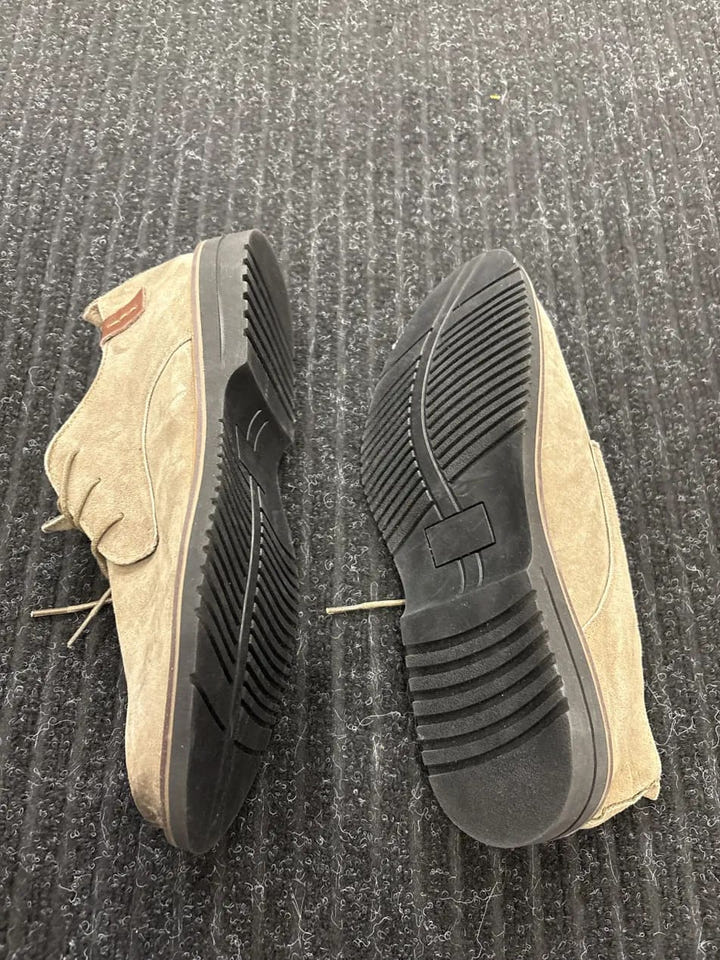 A pair of beige suede lace-up shoes shown from the side and bottom, revealing a black treaded sole, placed on a dark gray textured carpet.