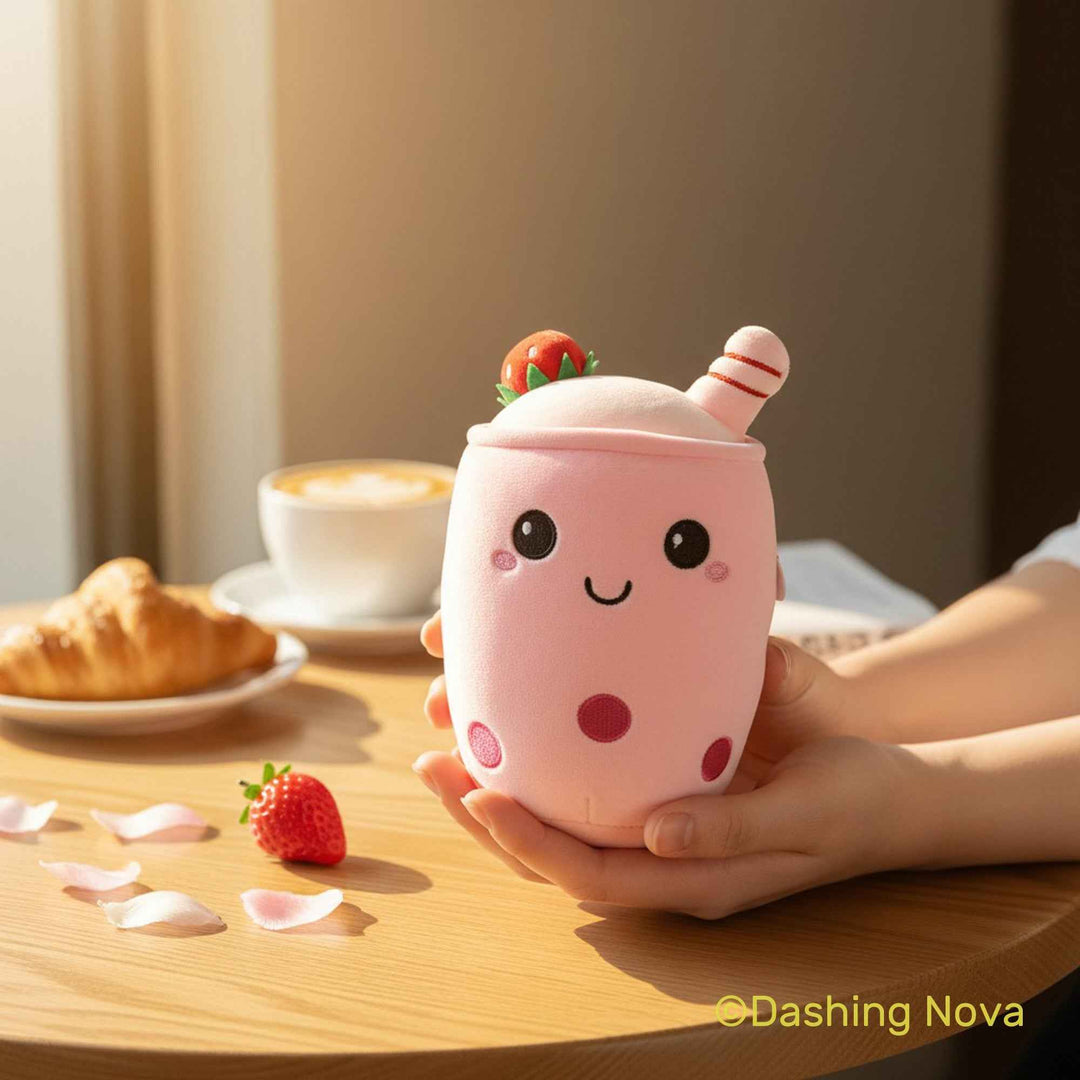 A cute pink boba milk tea cup character toy being held in hands over a wooden table, with a cup of coffee, a croissant, and a strawberry in the background.