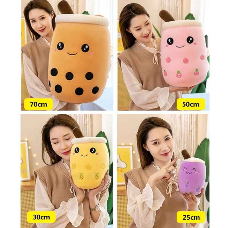 A 2x2 collage showing a woman holding four different boba-tube-shaped plush toys in different sizes: 70cm, 50cm, 30cm, and 25cm. The plushies are orange with black dots, pink with strawberry details, yellow with lemon details, and purple with cherry details.