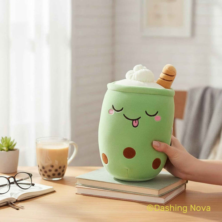 A green boba tea cup-shaped plush toy with a sleeping face, pink cheeks, and brown tapioca dots, topped with white foam and a striped straw, being held by a hand on a stack of books on a wooden table.
