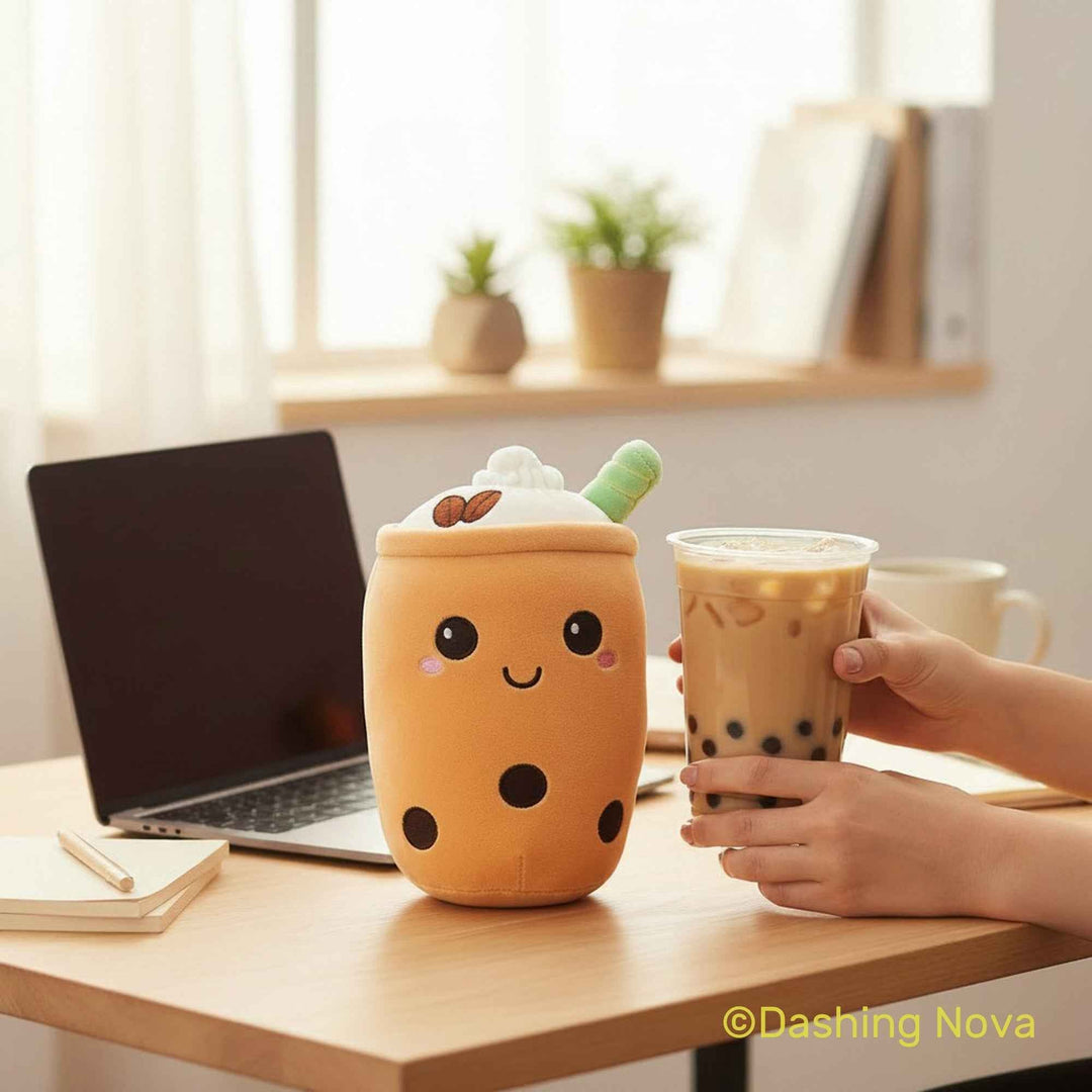 A cute orange boba drink plush toy with a smiling face, whipped cream top, and green straw sits on a wooden desk next to a laptop, with a hand holding a matching boba cup in front of it.