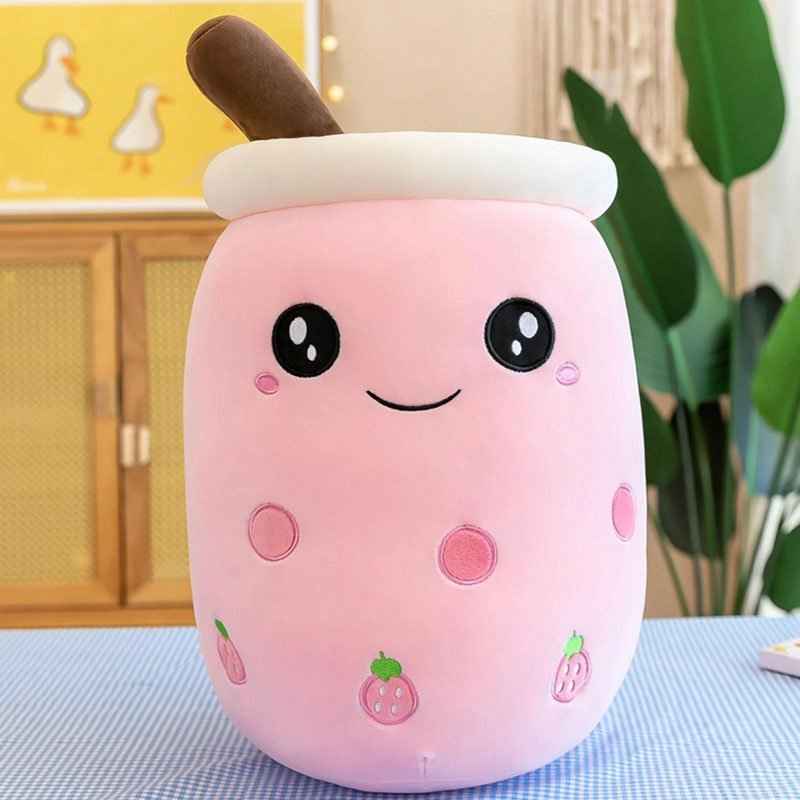 A pink plush toy shaped like a jar or cup with a brown handle, white rim, and embroidered face, set on a blue checkered surface in a softly lit indoor room with plants and wooden furniture in the background.